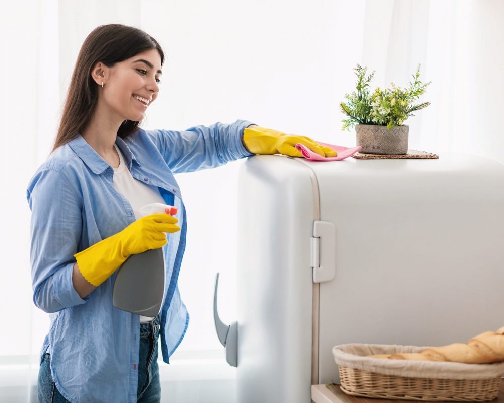 Cheerful young housewife cleaning fridge at kitchen Cleaning And Disinfection Concept. Smiling millennial woman in protective rubber gloves wiping white fridge surface standing at kitchen. Cheerful lady keeping her home tidy, using spray detergent